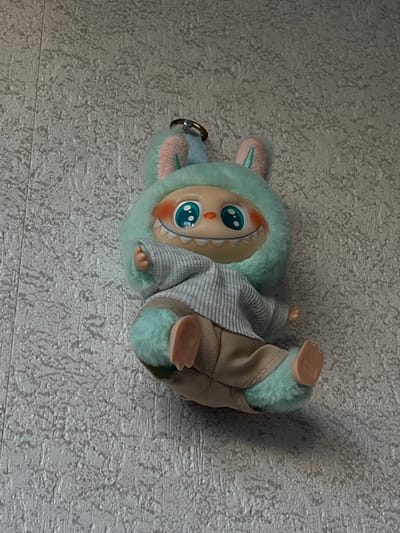 A plush toy with a light blue, fuzzy hood and pink bunny ears is attached to a grey, textured wall. The toy has a light beige face with large, blue, cartoonish eyes and rosy cheeks. It has a wide smile with visible white, jagged teeth. The toy wears a light grey, ribbed sweater and beige pants. Its arms and legs are light blue and fuzzy. The toy is positioned as if sitting or lying down, with its legs extended to the right. A metal keychain ring is attached to the top of its hood. The background is a plain, textured wall, typical of interior walls. The image was taken in Genemuiden, Netherlands.