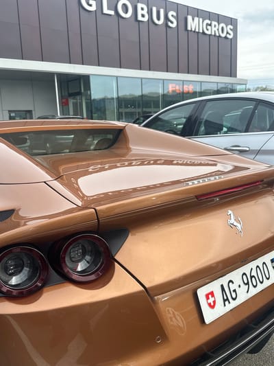 The image captures the rear-right section of a luxurious bronze-colored sports car, likely a Ferrari given the distinctive prancing horse emblem on its trunk. The car features prominent circular taillights, a slender red light strip, and a Swiss license plate displaying "AG 9600" alongside the Swiss flag. The vehicle's highly polished surface reflects its surroundings clearly.
In the background, a large commercial building stands, identified by the visible white letters "GLOBUS MIGROS" on its dark facade. Another business name, "Fust", can be seen on a window of the building and is also reflected on the car's body. The reflections on the car's surface are inverted and distorted, showing portions of the building's architecture and signage. Part of another, lighter-colored vehicle is visible to the left of the sports car.
The scene takes place outdoors during the daytime, under what appears to be an overcast sky, which provides soft, even lighting. The Swiss license plate and the prominent "GLOBUS MIGROS" and "Fust" signs firmly place the location in Switzerland, aligning with the provided context of Wallisellen. The overall image has a vertical orientation.