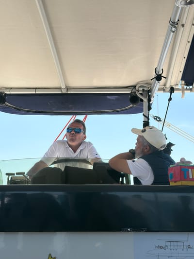 The image shows two men on a boat or yacht. One man, wearing a white baseball cap and with a white beard, is looking down with his hand near his face. Another man is steering the boat. A dark blue structure and white canvas are visible. It is not possible to determine from the image whether the location is Caniço, Portugal.