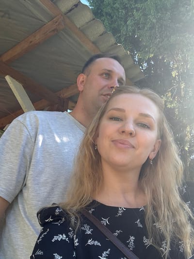 The image portrays two individuals, one female and one male, in an outdoor setting in Urmanets, Russia. The female, positioned in the foreground, appears to be in her late 20s to early 30s, characterized by long blonde hair, a light complexion with visible freckles, and a direct gaze towards the viewer with a slight smile. She is wearing a dark dress adorned with a white floral pattern and small hoop earrings. A dark strap, likely from a bag, crosses her chest. Behind and to the left of the female, a male individual, appearing to be in his late 30s to early 40s, with short dark hair and a light complexion, looks upward and to his left with a subtle smile. He is wearing a light grey short-sleeved t-shirt. The background on the left features a wooden support structure and a section of a corrugated roof. The background on the right is dominated by dense green tree foliage, with bright sunlight visible, producing a prominent lens flare on the right side of the frame.