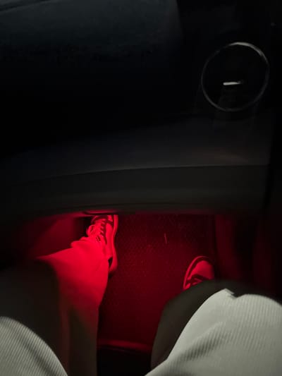 Inside a vehicle, a person's leg is visible, illuminated in red light. The leg is clad in white clothing and a sneaker with visible laces and a side stripe. The foot rests on a textured floor mat, also bathed in red light. The surrounding interior of the car is dark, with a round vent and a car seat partially visible.