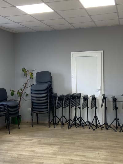 The image depicts an indoor scene, likely a rehearsal or meeting room, located in the city of Soroca, Moldova. The room has light gray walls and a suspended white tiled ceiling with a fluorescent light panel illuminating the space. In the center of the room, a stack of dark gray chairs is visible to the left, and to the right, a row of black music stands with perforated trays are set up. A white door with a silver handle and lock is situated in the background on the right. A small potted plant with sparse green and yellow leaves stands near the stacked chairs. The floor is covered with light-colored laminate flooring. The overall impression is one of a functional, sparsely furnished space.