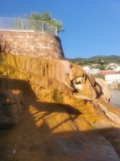 This media file depicts a natural hot spring area in Loutra Edipsou, Greece. The foreground is dominated by striking, weathered rock formations with a vibrant orange and yellow hue, likely colored by mineral deposits. Water appears to be flowing or present in pools at the base of these formations, creating a glistening effect on the rock surfaces. The texture of the rocks is rough and layered, suggesting significant erosion over time.
In the mid-ground, a brick wall curves around, topped with a metal railing and lush green foliage, suggesting a developed area or pathway near the natural springs. Beyond this, a hillside rises, covered in dense green vegetation. Nestled on the hillside is a cluster of buildings with white and red facades, characteristic of a town or village. The sky is a clear, bright blue, indicating a sunny day.
A prominent shadow of a person and a railing is cast across the mineral-rich rocks in the foreground, implying that the photo was taken by someone standing near the formations, possibly on a raised platform or walkway. The overall scene conveys a sense of natural beauty, geological interest, and a popular tourist destination where nature and human development coexist. The clear sky and bright sunlight suggest it is daytime. There is no visible text in the image.