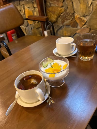 This image depicts a table laden with beverages and a dessert inside what appears to be a cafe or restaurant in Buôn Ma Thuột, Vietnam. The scene is observed from a slightly elevated perspective, looking down at a polished dark wooden table.
In the foreground, a white teacup with a gold rim sits on a white saucer. It contains a dark reddish-brown liquid, likely tea, with several reddish-brown fruits (possibly jujubes) and a white tea bag string submerged. A silver spoon rests on the saucer to the left. To the right of the teacup, a clear glass dessert goblet holds a white creamy substance, presumably yogurt or panna cotta, topped with diced yellow mango, a thin slice of lime, and a small sprig of rosemary. A decorative gold-colored spoon with a leaf-shaped handle is positioned to the right of the goblet.
Further back on the table, to the right of the dessert, a white coffee mug on a white saucer contains a light brown liquid, consistent with coffee with milk. A silver spoon is visible on its saucer. Adjacent to the coffee mug, a clear glass contains a dark amber liquid, possibly iced tea or another beverage.
The background features a rustic stone-clad wall composed of irregularly shaped stones in varying shades of brown, grey, and ochre. To the far left, a brown leather-like upholstered chair with a black metal frame and wooden armrests is partially visible, suggesting a seating arrangement. A red fire extinguisher is faintly discernible behind the chair. No individuals are visible within the frame.