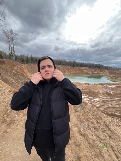 The image shows a person standing on a reddish-brown terrain with a body of water and a forest visible in the background. The person is wearing a black hooded jacket and has their hands near their face. The sky is cloudy and overcast. The location is likely Ivlevo, Russia, given the context. The landscape appears to be a former industrial site or a quarry, given the color and texture of the land and the presence of a body of water that may be artificially created. The image is oriented sideways.