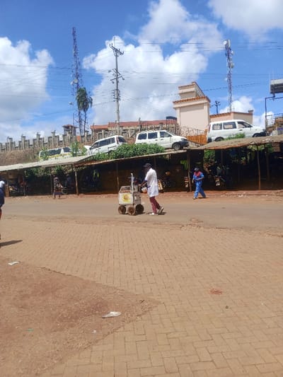 The image depicts a street scene in Kangema, Kenya, under a partially cloudy blue sky. In the foreground, a light brown brick-paved sidewalk transitions to a dirt patch on the left. The midground features an asphalt road where two individuals are visible. On the left side of the road, a man in a white shirt, dark trousers, and a dark baseball cap pushes a four-wheeled cart, which appears to contain items for sale. Further to the right on the road, another person wearing a blue jumpsuit walks leftward.

Along the immediate left edge of the road, a series of makeshift market stalls or covered structures with corrugated roofs are present, appearing to house small businesses. Obscured figures and parked motorcycles are visible within and behind these structures.

In the background, an elevated area is present, featuring a concrete retaining wall topped with a fence. Behind this fence, several white vehicles, resembling SUVs or vans, are parked. Further back, buildings with light-colored facades are visible. Dominating the upper background are two tall structures: a prominent blue lattice telecommunications tower with multiple antennas, partially obscuring a palm tree, and a utility pole with multiple power lines to its right.