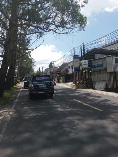 The image presents an outdoor scene in what appears to be a city street. On the road, a black Toyota Kijang car, bearing license plate "F 1443 KD," is moving away from the camera. The vehicle has some stickers at the back. Another vehicle is further ahead, though its details are less clear. The road is marked with a yellow line. The left side of the street is lined with tall trees, casting shadows on the road. On the right side, there are buildings, including one with a signboard displaying "BBRI ATM" and another with "HANIZA" advertising. Power lines and poles crisscross the scene above the buildings. The sky is visible above the buildings and trees, showing patches of blue and clouds. The location is labeled as Kioumaknao 2, Laos. There is no indication of actions or interactions of people in the scene.