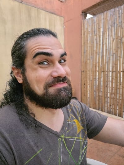 A forensic description of the scene in Villa San Andrés, Argentina, reveals an adult male captured in a mid-shot, presenting a direct gaze with a playful or mischievous facial expression. The individual has dark, long, curly hair pulled back, with a prominent beard and mustache containing visible grey hairs. His eyes are light-colored, possibly green or hazel, and his lips are slightly pursed into a subtle smile or smirk. He is wearing a dark grey, V-neck t-shirt adorned with an abstract graphic design featuring yellow and light green lines, along with a larger yellow crescent shape on the right shoulder. The background consists of a light tan wall on the left, and an orange-red wall above a vertical bamboo screen on the right. A tiled floor, possibly terracotta, is partially visible in the bottom right corner.