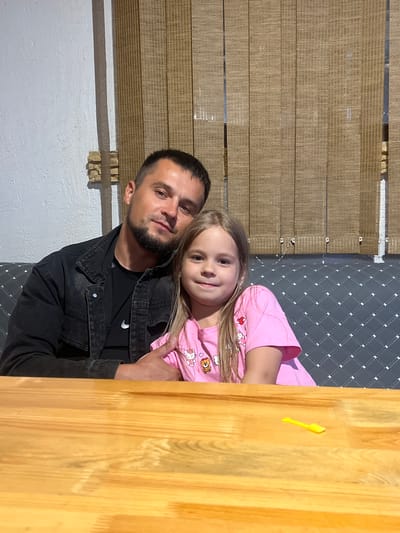 In this image, a man and a young girl are posing for a photo. The man, with a beard and short hair, wears a dark denim jacket over a black t-shirt. The girl, with blonde hair, is wearing a pink t-shirt with cartoon characters. She is leaning her head on the man's shoulder. The background features a textured wall, a woven material, and a wooden surface. The lighting suggests it is daytime, with natural light illuminating the scene. A small yellow object is visible on the wooden surface. The overall mood appears affectionate and familial. No text is visible in the image.