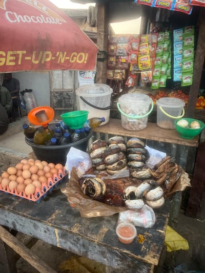 A street market stall is packed with diverse goods. Shelves are stocked with sachets of spices and snacks. Clear buckets contain peanuts, onions, and tomatoes. A black pail holds bottles of oil and green containers. A tray of brown eggs rests beside the pail. Smoked fish and some sealed bags are placed on a weathered wooden surface. A sign with "Chocolate" and "GET-UP-'N-GO!" is visible.