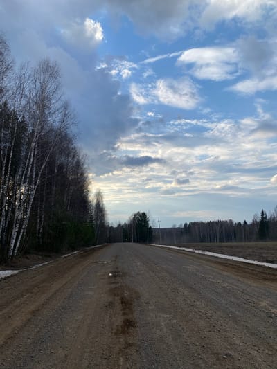 The image shows a dirt road running through a rural area. The sky is filled with striated clouds. A line of trees borders the road on both sides. Some snow is visible along the road edge. Power lines are visible in the background. The location is claimed to be in Nekrasovo, Russia. There are no people visible.