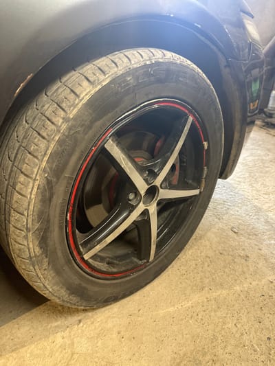In the city of International, Kyrgyzstan, a close-up, eye-level shot captures the front passenger-side wheel of a dark-colored car, possibly a sedan, resting on a concrete floor. The tire is a Rotational Radial tubeless tire with the size marking "215/55R17 98W" visible. The wheel is a black alloy rim with silver accents and a red stripe around its circumference. The tire itself appears dusty and moderately worn. Part of the car's fender and bodywork is visible in the upper left and right portions of the frame, with the background showing a dimly lit, possibly industrial or garage-like environment. The lighting suggests the image was taken indoors, with artificial light casting reflections on the car's paint and the concrete floor. No people or other vehicles are present in the frame.