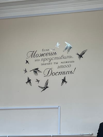 The scene depicts an interior wall within a structure in Dmitrievskaya, Russia. The wall is a solid beige-off-white color, topped by a white crown molding along the ceiling line. Centrally positioned on the wall is a large, black, stylized wall decal featuring Russian text and eleven bird silhouettes in flight. The Russian text reads, "Если ты Можешь это представить, значит ты можешь этого Достичь!", which translates to "If you can imagine it, then you can achieve it!". The bird silhouettes are depicted in various sizes and orientations, appearing to fly upwards and outwards from the text, with some having a lighter or reflective appearance. In the lower portion of the image, a white horizontal railing-like structure is visible, extending across the frame. Below this structure, a white pipe or conduit runs horizontally along the wall. The lower right corner of the image captures a partial view of a person's upper back and shoulder, dressed in dark attire, facing away from the camera.