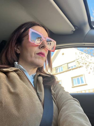 The image displays a female individual seated in the front passenger or driver's seat of a vehicle, looking towards the right. She has dark brown hair and is wearing oversized, square-framed sunglasses with prominent pink-tinted lenses, which reflect the upper section of a light-colored building. Her lips are covered in red lipstick. She wears a light blue striped collared shirt under an unzipped tan or light brown suede-like jacket. A dark-colored seatbelt is fastened across her chest and left shoulder. The vehicle's interior features a light-colored headliner and a partial view of a sunroof opening. Outside the vehicle, a multi-story building with light-colored walls, stone detailing around its upper windows, and several windows fitted with white roll-down shutters is visible against a clear blue sky. Bare tree branches are present on the right side of the exterior view. The scene is located in the city of Roodt-sur-Syre, Luxembourg.