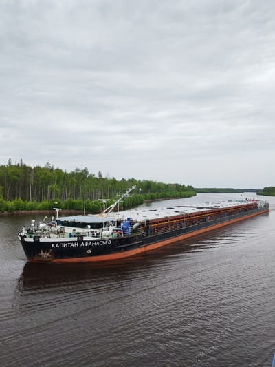 A large, long barge, identified by the Cyrillic text "КАПИТАН АФАНАСЬЕВ" (Captain Afanasyev) on its bow, navigates a wide river. The barge is laden with what appears to be cargo, covered by a tarp and supported by railings, with several orange and yellow structures visible along its length. The vessel has a black hull with an orange-red waterline. In the foreground, the dark water shows ripples and wake from the barge's movement. The background features a dense, green forest lining the riverbanks, with trees of varying heights and lush foliage. The sky above is overcast with a grey, cloudy, and diffused light, suggesting a temperate or cool climate. The scene depicts the barge actively transporting goods through a natural waterway, likely in a rural or undeveloped area, given the thick forestation. The overall mood is serene and functional, emphasizing the commercial activity against a backdrop of natural scenery. The time of day appears to be daytime, given the visibility. The location is identified as Kurdiug, Russia.