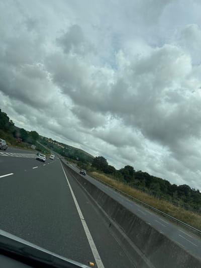 The image shows a view from a vehicle traveling on a highway in the New Ross Municipal District, Ireland. The highway has multiple lanes separated by white lines, and a concrete barrier runs along the right side. Several cars and a caravan are visible on the road. The surrounding environment consists of rolling green hills covered in trees and grass. The sky is overcast with dramatic, heavy clouds. The overall impression is a cloudy day on a scenic highway.