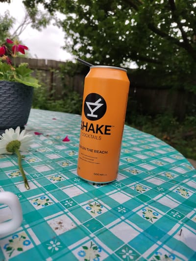 A medium shot captures an orange aluminum can of "SHAKE Cocktails" with a black straw inserted, resting on a green and white checkered tablecloth. The can features a stylized white logo and the text "SHAKE COCKtails" prominently displayed. Below, smaller text reads "SEXX ON THE BEACH" followed by Cyrillic script. The tablecloth is adorned with a floral pattern of blue flowers and green leaves. To the left of the can, a white daisy with a green stem lies on the tablecloth. In the background, a dark gray planter holds vibrant pink flowers and green foliage. Beyond that, a wooden fence is visible, and further in the distance, lush green trees and a cloudy sky form the backdrop. The scene appears to be outdoors, likely in a backyard or garden setting in Okhtyrka, Ukraine.