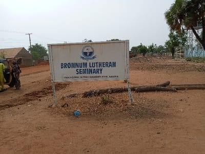 The image shows a sign in front of a building, located in Rumde Waziri, Nigeria at the coordinates provided. The sign is white with blue lettering, and it reads "BRÖNNUM LUTHERAN SEMINARY, YOLA P.O. BOX 74 YOLA ADAMAWA STATE, NIGERIA." A large tree can be seen to the right of the sign. The ground is dirt and there is a small blue object in the foreground. Two people appear to be walking away from the camera in the distance. The overall scene is that of a building entrance in a rural location.