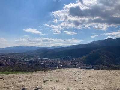 The image presents a wide, daytime view overlooking a cityscape nestled within a valley in Sveti Konstantin, Bulgaria. The foreground shows a barren, rocky landscape transitioning into a vantage point. Below, the city sprawls with tightly packed buildings and infrastructure, including what appears to be a white industrial complex. The surrounding valley walls rise into dark, forested mountains in the midground. The sky is a light blue with scattered, fluffy white clouds. There are no visible people, subjects, actions or interactions.