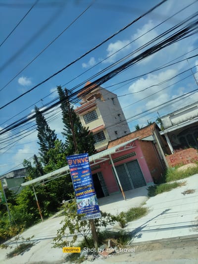 In Gò Dầu, Vietnam, a street scene unfolds under a bright, cloudy sky with numerous power lines crisscrossing the frame. In the foreground, a blue banner displays Vietnamese text, advertising a service or event. Behind it, a pink building with a red-tiled roof and metal gates stands next to a taller, cream-colored building with a terracotta roof. Lush green trees and vegetation border the sidewalk and street. The overall impression is a typical urban or suburban landscape in Vietnam, characterized by its infrastructure and plant life.