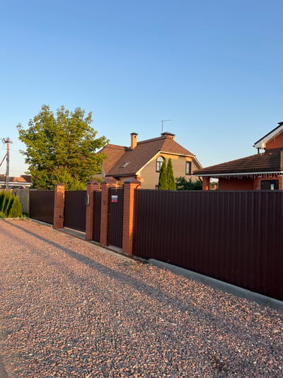The image shows a view of houses and a fence with a gravel-covered ground in front of it, taken from a side angle. The sky is clear and blue, suggesting fair weather. The houses appear to be suburban residences, with architectural details like tiled roofs, brick facades, and chimneys. A large tree with green foliage is visible, adding to the residential setting. The fence is made of corrugated metal and brick pillars. The ground is covered in small, dark-colored gravel.
The lighting indicates that it is either early morning or late afternoon, as the sun casts long shadows. The scene appears to be in a quiet residential area, likely a street or driveway. There is no specific activity or event depicted; it is a still scene of a residential property.
Notable details include the textured surface of the gravel, the varying shades of brown in the fence and roofs, and the bright green of the tree. A small sign with red and blue lettering is attached to the fence near a gate. The overall impression is one of a peaceful, well-maintained property on a clear day. The text on the sign is not legible.