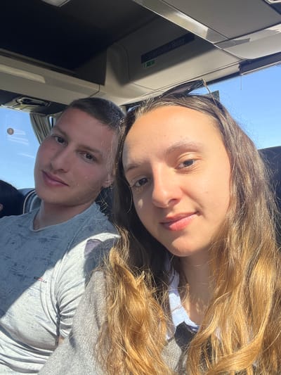 The image depicts two young adult individuals inside a Mercedes-Benz coach or bus, located in Otero de Herreros, Spain. The female on the right, with long light brown hair, wears a light collared shirt under a grey knitted garment and has black-framed sunglasses perched on her head. She is holding the camera, taking a selfie. The male on the left, with short dark hair, wears a light grey patterned short-sleeved t-shirt with a partially visible red logo. Both individuals are looking towards the camera with slight smiles. Bright daylight enters from the right, casting shadows on the male's shirt and face. A clear blue sky is visible through the window on the left side of the vehicle. The interior includes a light-colored overhead compartment with a "Mercedes-Benz" label and a green indicator light.