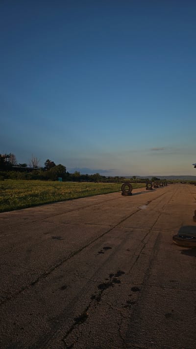 This image captures an outdoor scene in Buhovtsi, Bulgaria, at what appears to be an aged, concrete or asphalt runway or wide path, extending into the distance under a late afternoon or early evening sky.
The upper approximately one-third of the image consists of a clear, deep blue sky, transitioning to a lighter, more yellowish hue near the horizon on the right. Several minute, dark specks are visible against the blue sky.
The mid-ground features a broad expanse of green, unkempt grass or vegetation running parallel to the paved surface on the left. Within this grassy area, slightly to the left of the image center, a light blue portable toilet is visible. Further back on the left, a cluster of trees, some with bare branches, partially obscures a structure or building with what appear to be antennae or communication equipment. Distant hills and faint cloud formations are visible along the horizon line on the right.
The lower two-thirds of the image depict a dark, cracked concrete or asphalt surface. This surface shows numerous fissures, lighter patches, and dark, irregular stains. A series of approximately eight dark, worn tires are positioned upright at intervals along the right edge of this paved surface, extending from the mid-foreground into the distant background. A portion of another tire is visible in the extreme lower right foreground. Long, dark shadows are cast across the paved surface from the right, indicating a low-angle light source. No individuals are present, and no discernible actions or interactions are occurring.