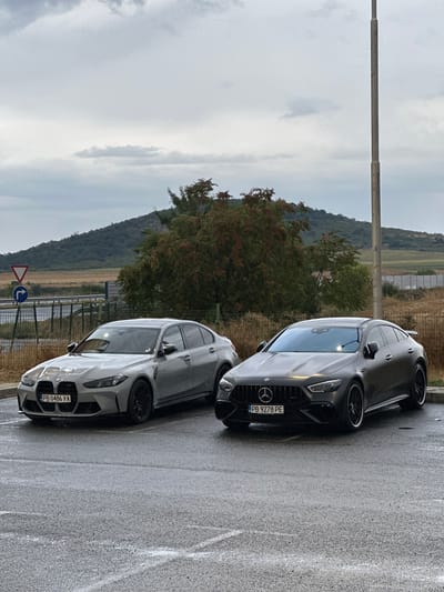 Two high-performance cars, a grey BMW M3 Sedan and a dark grey Mercedes-AMG GT 63 S 4MATIC+ 4-door coupe, are parked on a wet asphalt surface in Drazhevo, Bulgaria. The BMW, license plate PB 0486 YA, is on the left, and the Mercedes, license plate PB 9278 PE, is on the right. The background features a hill covered in vegetation, a road, and a cloudy sky. A lamppost stands to the right of the vehicles. The scene appears to be a parking area, possibly near a roadside or a rest stop.