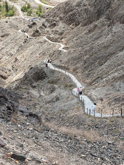 The image depicts a winding paved pathway descending a rocky, arid mountainside. The path is lined with a metal chain railing supported by wooden posts. Several individuals are visible on the pathway. In the foreground, a person wearing a light-colored robe walks down the path. Further up, a group of three people, two wearing hats and one in a patterned dress, walk in the same direction. There are red signs with Chinese characters placed along the path. The surrounding environment is characterized by sparse vegetation, dry grasses, and loose stones and rocks. The terrain is steep and appears to be a natural canyon or gorge. The sky is not visible. The pathway appears to be a tourist trail, possibly leading to or from an attraction. It is likely located in a mountainous or desert region of China, given the presence of Chinese signage and the arid landscape. The context of Jiayuguan, China, suggests this might be a section of the Great Wall or a related historical site, known for its mountainous terrain and historical fortifications.