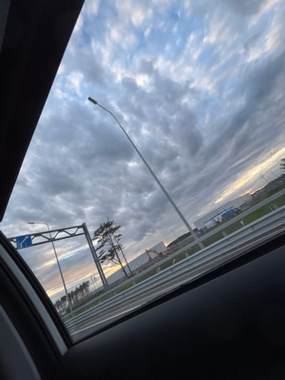 The image, taken from inside a vehicle with an open sunroof, shows a view of the sky and the exterior environment.  The sky is filled with textured clouds, illuminated by a setting sun.  A tall streetlight extends upwards. A road sign is visible on the right. A line of trees appears in the distance, adjacent to a fenced off area. The sunroof's edge and surrounding vehicle interior are visible in the foreground. The scene is in Levashovo, Russia.