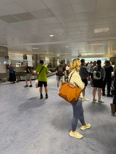 This image captures an indoor scene at what appears to be an airport terminal in Granadilla de Abona, Spain, given the provided context. Numerous people, predominantly adults, are visible standing in queues or waiting areas. Most individuals are dressed in casual travel attire, including t-shirts, shorts, leggings, and sneakers, and many are carrying luggage such as backpacks and shoulder bags.
In the foreground, a woman with blonde hair, wearing a light top and blue leggings, carries a prominent large orange tote bag. Behind her, other travelers are also waiting, some standing near what appears to be a baggage claim carousel or check-in counter structure. The floor is a light blue, mottled surface, and the ceiling is composed of white grid panels with various integrated lights, including fluorescent strips and circular fixtures.
The activity depicted is people waiting, consistent with an airport environment where passengers queue for check-in, security, or to collect luggage. The atmosphere appears to be orderly, with people patiently standing in line. No strong emotions are overtly displayed. Visible text is limited; a black sign with light text, possibly "DHL," can be faintly seen on a piece of equipment in the mid-ground. The lighting suggests a bright indoor setting, likely during the day.