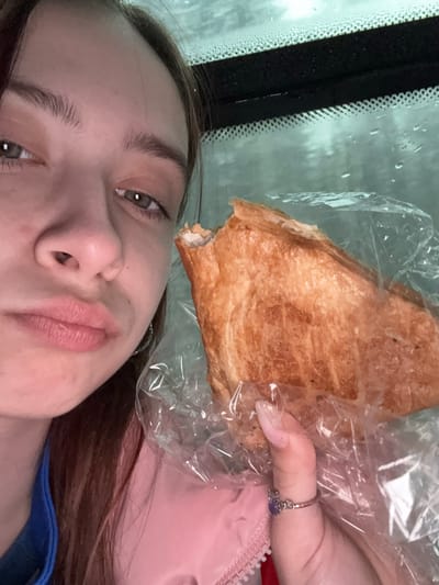 A young female is holding a pastry inside a clear plastic bag, likely within a vehicle due to the visible window and condensation. The specific location of Lunezhki, Russia is not discernible in the image. No actions or interactions are apparent aside from the subject holding the pastry. The environment appears to be indoors or inside a vehicle, given the window and perceived interior setting.