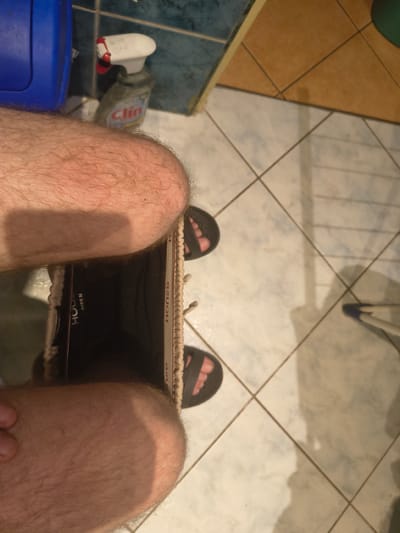 The image depicts a person's legs and feet from a low, Dutch angle perspective, showing hairy skin and feet wearing black flip-flops. The person is positioned in what appears to be a bathroom or utility area. In the background, a blue plastic container is visible to the left, and a spray bottle labeled "Clin" with a red nozzle is to its right, positioned against a tiled wall. The floor is tiled with a pattern of light-colored tiles with dark grout lines. The scene suggests a domestic environment, possibly in Grójec, Poland, given the context of the request.