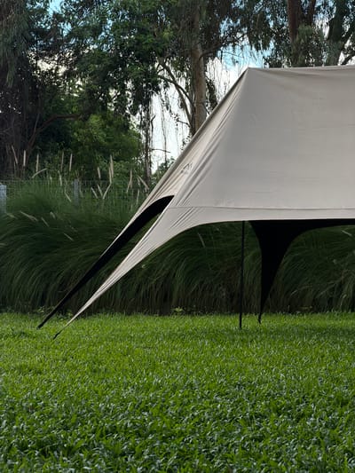A light-colored, angular tarp shelter is set up in a grassy area with tall green grass and a fence in the background. The shelter has several poles supporting it, and one side appears to have a dark opening or cutout. Trees with green leaves and some dry branches are visible behind the shelter and fence. The sky is partly cloudy. The scene is outdoors and appears to be in a park or a garden. The location is Tambon San Phi Suea, Thailand. There are no people visible in the image.
