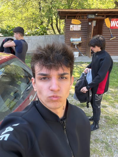 The scene shows three males outdoors. Two are dressed in wetsuits, with one appearing to pack a backpack near a small wooden building, while the other stands closer to the camera. The third person is partially visible behind a red car. The setting appears to be a natural area near a wooden structure. The exact location within Poleto, Bulgaria, is not discernible from the image.