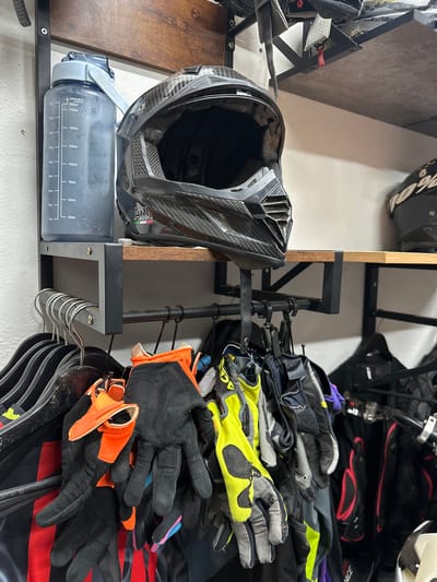 The image shows a close-up, eye-level, medium shot of motorcycle gear storage. In the background, a wooden shelf holds a black motorcycle helmet with a carbon fiber pattern, and another black helmet is partially visible on the right. Below the shelf, a rack is filled with hangers holding various pieces of motorcycle apparel, including multiple pairs of gloves in different colors (black, orange, neon yellow) and some dark-colored garments. A large, gray water bottle with measurement markings is attached to the wall on the left, next to the helmet. The scene appears to be indoors, possibly a garage or storage area, with white walls and a dark metal support structure for the shelf and rack. There are no people visible in the image. The location of Dedovsk, Russia, is not depicted or implied by any visual elements in the image.