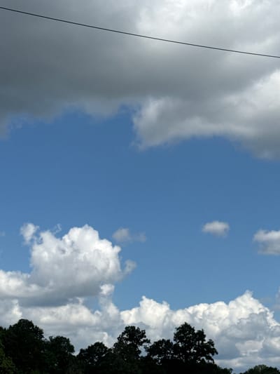 The image displays a serene outdoor scene with a clear blue sky dominating the upper portion. Several white, fluffy clouds are scattered across the sky, with some larger, darker clouds towards the top. A thin, dark wire stretches horizontally across the sky in the upper part of the frame. The bottom of the image features the dark green silhouette of a treeline, suggesting a natural environment. The scene appears to be set in Wysiedle, Poland, likely a rural or suburban area given the presence of trees and open sky. There are no people, objects, or actions depicted that would suggest a specific event or narrative. The overall impression is one of natural beauty and tranquility under a partly cloudy sky.