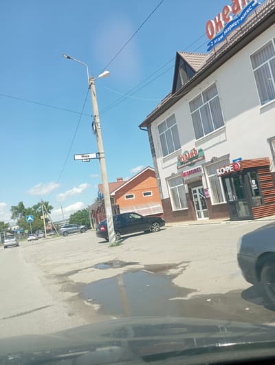The image depicts a street scene in the city of Otradnaya, Russia, under a clear blue sky with scattered clouds. A concrete lamppost stands prominently in the foreground, with electrical wires running across it and into the sky. A street sign attached to the lamppost reads "Мира" (Mira) with numbers "8-17" below it, indicating a street name and house number range.
To the right of the lamppost is a white building with a dark brown lower facade and a brown-tiled roof. A large red and blue sign on the roof reads "Океан" (Ocean) with smaller text below it in blue that says "2 этаж Маркет-микс" (2nd floor Market-mix). On the white facade of this building, there are several large windows with white frames and light-colored curtains. Below some of the windows are signs for businesses: one in green text says "КУРАЖ" (Kurazh) with additional text below in smaller font "ОХОТА-РЫБАЛКА-ОДЕЖДА-СПОРТ" (Hunting-Fishing-Clothing-Sport). Below that, a sign reads "wildberries". To the right of the "wildberries" entrance, there is a small kiosk-like structure with a sign above it that says "КОФЕ 24" (Coffee 24) and displays a red "24" icon.
In front of the building, a black car is parked. Further down the street, a black sedan is visible. Other cars are present in the background, including a silver SUV and a silver hatchback. A traffic sign with a blue background and white symbols is visible on the left side of the road. The road surface is asphalt with several large puddles and some scattered debris. Trees and other buildings are visible in the distance, suggesting an urban or semi-urban environment. The image appears to have been taken from inside a vehicle, as the bottom of the frame shows the reflection on a car's dashboard or windshield.