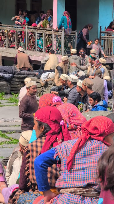 The image depicts a gathering of people in Malana, India, likely during a festival or community event. The majority of individuals are seated or standing on a stone pathway and a raised platform. Many are wearing traditional Himachali caps, a distinct white conical hat. Several individuals are dressed in woolen shawls and jackets, indicative of the mountain climate. In the foreground, a man in a brown sweater and a white cap is looking towards the right, engaged in conversation or observation. Behind him, several people are seated on the ground. Women are visible wearing colorful headscarves and traditional clothing. On a higher level, a group of people, including children, are standing and sitting on a balcony or terrace, looking down. The architecture suggests a rural mountain village setting. The environment appears to be outdoors with natural stone and rough-hewn structures. The overall atmosphere seems to be one of a communal gathering.