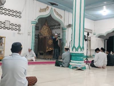 The interior of a mosque in Tumpeun, Indonesia, is depicted, showing a religious gathering. A male speaker stands center-right on a raised pink platform, wearing a black shirt, patterned sarong, and white peci, holding a microphone. Behind him is an intricately carved wooden mihrab or mimbar adorned with golden Arabic calligraphy. To the speaker's right, a tall, striped white and teal column is visible, with a black microphone stand positioned nearby.
Approximately seven to ten men are seated on the white tiled floor, facing the speaker. In the foreground, a man with his back to the viewer wears a light blue shirt, plaid sarong, and dark peci. To the left of the speaker, another man in a white robe and white peci sits on the floor. To the right of the speaker, another man in a grey shirt, green sarong, and white peci is seated, with a clear glass of water on the floor in front of him. Further right, a group of three additional men, one in a white patterned shirt and black peci, are also seated.
The mosque features white walls, some with dark patterned designs resembling bricks. Wall-mounted fans are visible. A simple wooden shelf filled with books is on the far left. On the far right, a whiteboard with visible writing is mounted on the wall. The ceiling is light blue with white trim, and artificial lighting illuminates the space. A small, open book on a wooden stand rests on the floor near the right-side column.