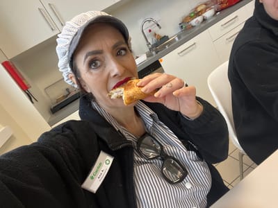 A woman identified as Ana A. from the "Cactus Poissonnerie" is eating a croissant in what appears to be a kitchen setting. She is wearing a striped shirt, a black jacket, glasses on her shirt, and a hairnet. There is a sink and kitchen counter visible in the background. The exact location in Mensdorf, Luxembourg, cannot be determined from the image.