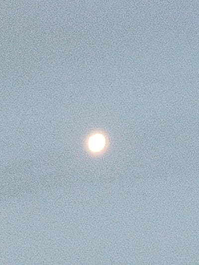 A pale, luminous orb occupies the center of the frame against a washed-out, light blue background. The texture of the backdrop suggests a hazy or overcast sky. The light source lacks sharp definition, appearing softly diffused. The surrounding expanse is uniform in color and texture, devoid of any distinct features or objects apart from the central point of light.