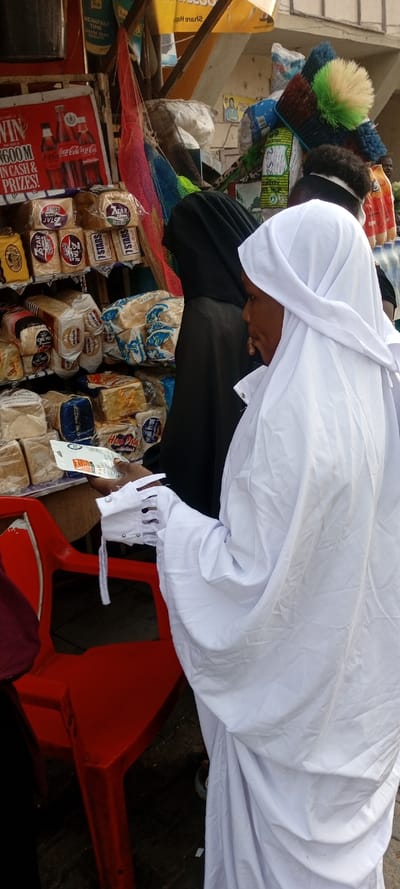 A woman in white, possibly religious attire, stands at a shop with a red chair nearby. The shop sells bread, with brands like "7 Stars" and "Home Pride" visible. A red Coca-Cola advertisement with the text "WIN 600M IN CASH & PRIZES!" is prominently displayed. Behind the woman, another individual in a black garment stands near brooms and mops. The scene appears to be a bustling market area.