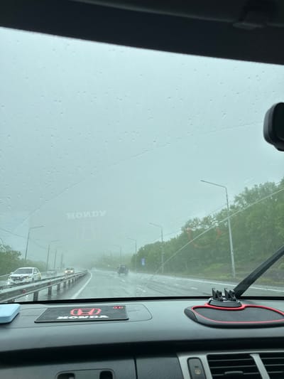 The image captures a view from inside a vehicle driving on a highway in Svetly, Russia, during rainy weather. Water droplets are visible on the windshield, obscuring the view of the road ahead. The highway is wet, and other vehicles are visible in the distance. On the dashboard, a red and black mat with the "HONDA" logo is present. The sky is overcast, contributing to the overall dim lighting. The surrounding environment appears to be a mix of trees and streetlights, indicating an urban or suburban setting. No people are clearly visible within the frame, apart from the implied driver of the vehicle.