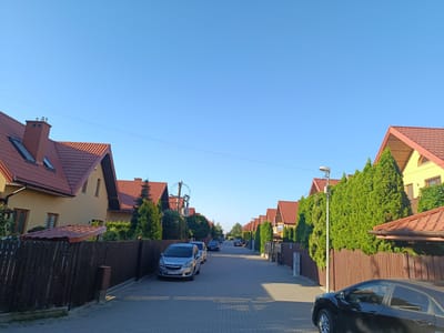 A wide, straight, paved street in Grójec, Poland, is lined on both sides with identical houses. The houses have yellow walls and red tiled roofs, with tall, manicured evergreen trees planted along the fences. Several cars are parked along the street, and a streetlamp stands on the right side. The sky is clear and blue.