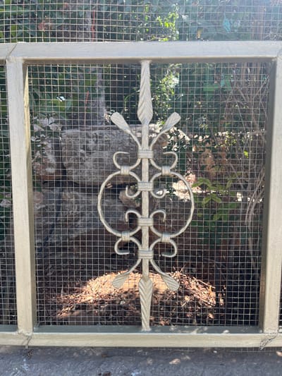 A section of a light-colored, possibly beige or pale green, metal fence panel is depicted. The panel features a rectangular frame with a fine, square-grid wire mesh infill. Centrally, an ornate vertical wrought-iron-style decorative element, painted to match the frame, displays scrollwork and stylized leaf motifs at its top, middle, and bottom. Behind the mesh, a dense natural environment is visible, comprising green foliage, tree branches, large grey-brown rock formations, and a ground cover of dried leaves and debris. Strong natural light illuminates the scene, casting shadows, particularly on the paved, light-colored ground surface directly in front of the fence panel. No individuals or animals are present. The precise location within the Menashe Regional Council, Israel, is not discernible from the visual information provided.