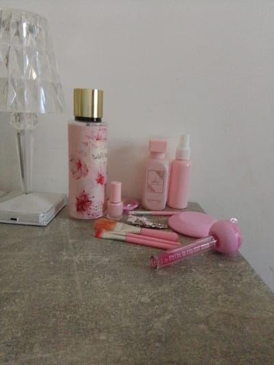 The image shows a collection of pink-themed cosmetic and beauty products arranged on a grey, speckled surface, likely a tabletop or dresser. To the left, a clear, faceted lamp with a diamond-like pattern stands next to a white rectangular object that appears to be a light or a charger. Dominating the center is a tall, pink bottle with a gold cap, decorated with floral patterns. Beside it, a small pink bottle of nail polish stands upright. Further to the right, two smaller pink bottles, one with a square cap and the other with a spray nozzle, are visible. In the foreground, several makeup brushes with pink handles and orange-pink bristles are laid out, along with a pink oval-shaped object and a clear tube with pink liquid and a pink ball on top. The background is a plain, light-colored wall. There are no people or actions depicted. The location is not specified within the image or any provided context.