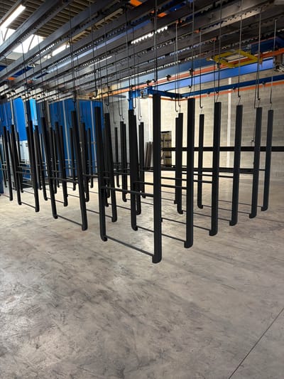 The image shows a production line in a factory setting in Mol, Belgium. The primary subjects are numerous black metal components, likely parts being manufactured or prepared for coating, suspended from an overhead conveyor system. These components are arranged in parallel rows and extend across the frame. The conveyor system itself is visible with its metallic tracks and supporting structures, colored blue and orange.
The scene appears to be an indoor industrial environment, with a concrete floor that shows signs of wear and tear, including scuff marks and cracks. Natural light or overhead lighting illuminates the space, casting shadows and highlighting the texture of the concrete. In the background, other factory equipment and structures are partially visible, suggesting an active manufacturing facility. There is no visible text in the image. The time of day and weather are not discernible. The overall impression is one of industrial efficiency and production.