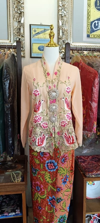 A detailed photograph captures a traditional Peranakan attire, consisting of a kebaya top and a batik sarong skirt, displayed on a headless white mannequin with a gold-colored neckpiece. The ensemble is showcased indoors, likely in a shop or cultural exhibition, with other garments and historical items visible in the background.
The kebaya top is a delicate light peach or beige color, featuring long sleeves. Its most striking feature is the elaborate embroidery on the front opening and collar, which depicts intricate floral motifs in shades of red, pink, and green, interspersed with stylized white and red birds resembling peacocks or phoenixes. The embroidery appears to be a form of eyelet or cutwork lace, adding texture and depth to the garment. A decorative, silver-toned kerongsang (brooch) with multiple sparkling segments fastens the front of the kebaya, adding a touch of elegance.
The sarong skirt is made of richly patterned batik fabric. The background color is a warm reddish-orange, adorned with large, vibrant floral designs in blue, pink, and green, along with swirling foliage. The patterns are dense and intricate, characteristic of traditional batik.
The background of the image reveals additional context. On the left side, a dark wooden display cabinet or shelf unit holds various items. On one shelf, several beaded and pearl necklaces are visible inside a clear container. Below, another item wrapped in plastic shows a hint of a blue pattern. Hanging next to the mannequin on the left are dark-colored garments (green, black) covered in clear plastic, suggesting they are also for sale or display. Part of a large, ornate silver-colored frame is visible on the upper left, complementing the traditional aesthetic.
In the center-top of the image, mounted on a light-colored wall, is a framed historical document. Text visible on this document includes "CHUNG THYE PHIN", "SHARE", and "10 cents", along with a date "1.1.1918". This appears to be an old share certificate.
On the right side, another large, ornate silver-colored frame, matching the one on the left, is partially visible. A second framed document or poster can be seen within this frame, bearing the text "IN KEE CHAN" and "TEN". To the right of the mannequin, red garments, also covered in plastic, hang on a rack, similar to the left side. A wooden display stand or shelf on the lower right holds folded fabric, possibly more batik, and a white enamel bowl with a floral pattern inside it.
The overall scene evokes a sense of cultural heritage and traditional craftsmanship, with the display hinting at the sale or exhibition of authentic Peranakan clothing and related artifacts.