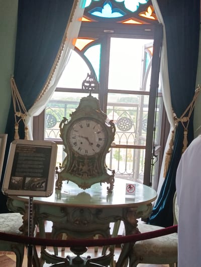 The image depicts an ornate, light green antique mantel clock placed on a matching decorative table. The clock features Roman numerals and intricate golden detailing, with the hands indicating approximately 10:10. The table is also elaborately carved and has a light green finish.
The setting appears to be indoors, possibly a museum or historic house, given the presence of display information and a velvet rope barrier. To the left of the clock and table, there is a framed display with text and images, suggesting an exhibit. A sign with a "do not touch" symbol (a hand with a red circle and slash) is visible on the table.
Behind the table and clock, a large window with stained glass accents in blue and orange is prominent. The window is partially open, revealing a glimpse of the outdoors with greenery and architectural elements, possibly a balcony or fence. Dark blue curtains with decorative tassels frame the window on either side.
A person is partially visible on the right side of the frame, seen from the back and shoulder, wearing a white garment. The overall ambiance suggests a preserved historical space, possibly a room within a palace or manor. The lighting suggests it is daytime, and the view through the window indicates fair weather. The scene is calm and focused on the display of the antique clock and its surroundings.